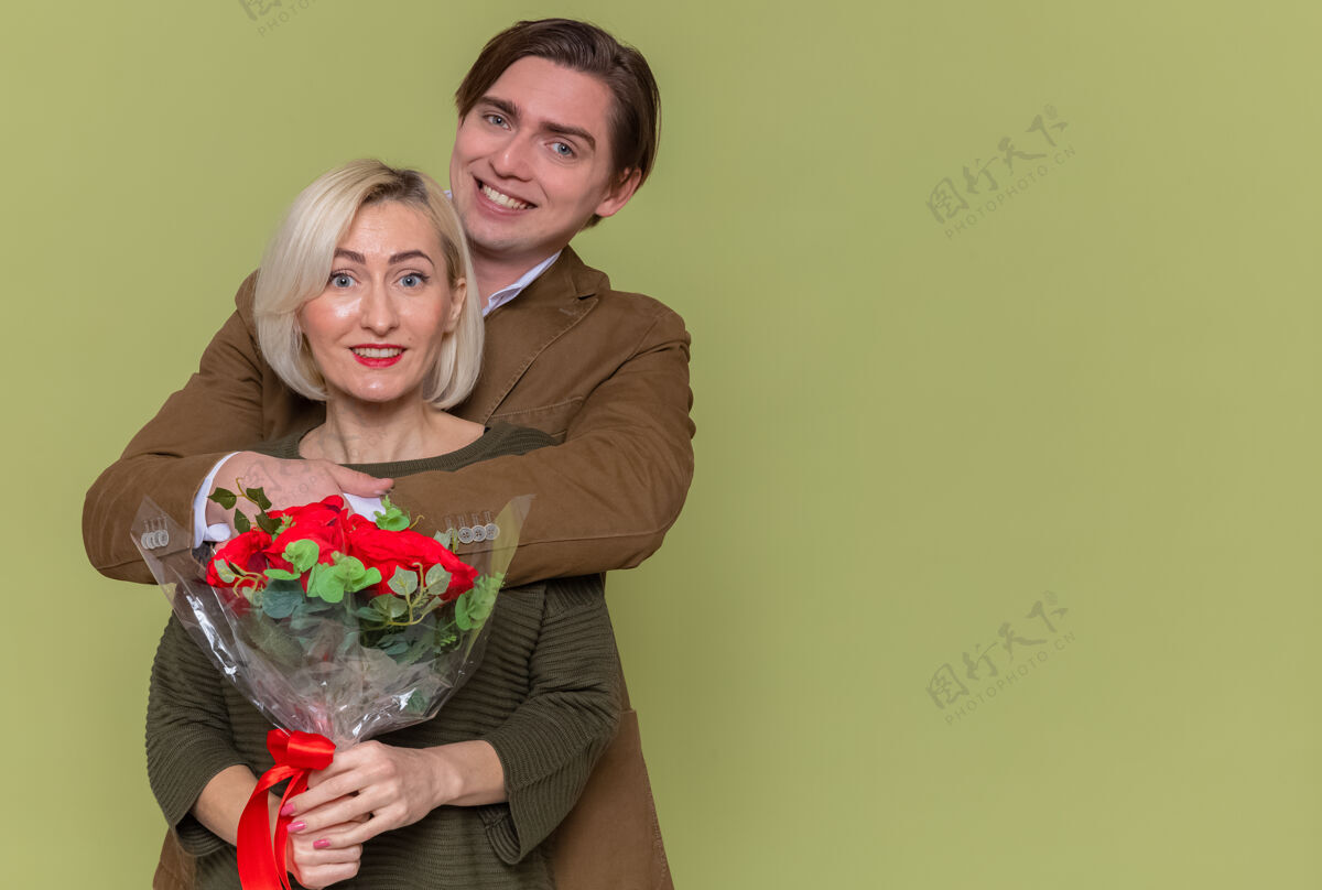 一起年轻美丽的夫妇幸福的男人捧着一束红玫瑰和女人拥抱着幸福的爱情一起庆祝国际妇女节站在绿色的墙上男人爱庆祝