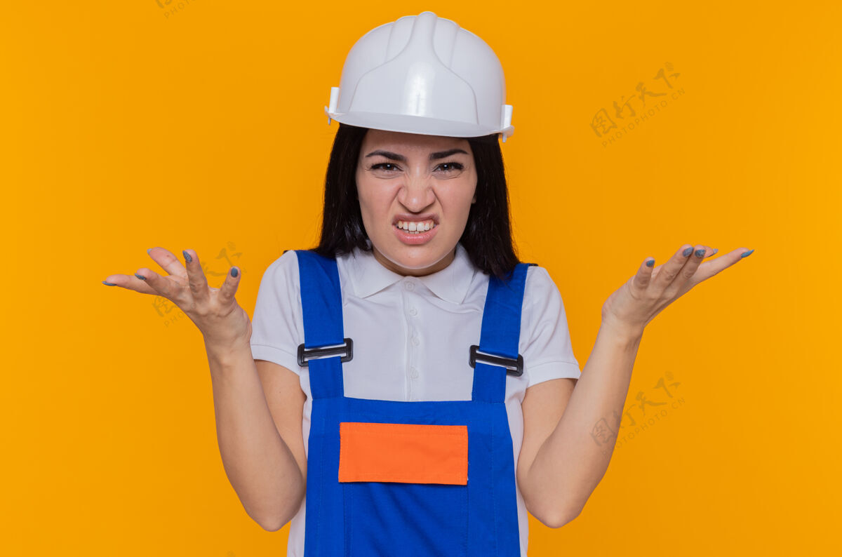 手臂身穿建筑制服 头戴安全帽的年轻建筑女工人站在橘色的墙上 看着前面恼怒地举起双臂站立恼怒建筑工人