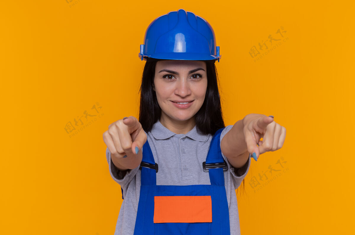 建筑身穿施工制服 头戴安全帽的年轻建筑工人自信地微笑着 双手食指指着前面站在橙色的墙上微笑建筑工自信