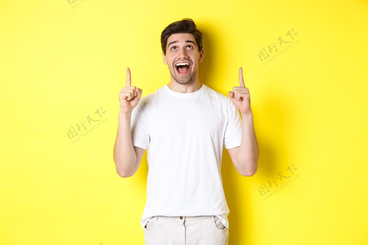 帅一个穿着白色t恤的快乐的年轻人对促销活动做出了反应 他惊奇地指着并抬起头 站在黄色的背景上时尚男人男人