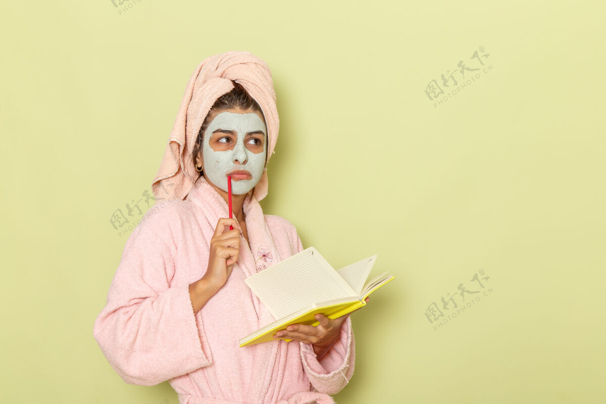 女士正面图穿着粉红色浴袍的年轻女性在绿色的表面写下思考的笔记思考漂亮笔记