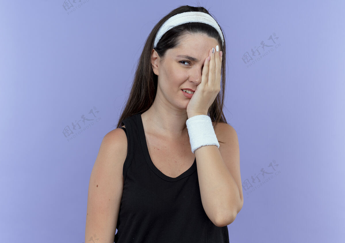 年轻愤愤不平的年轻女健身者戴着头巾 一边捂着一只眼睛 胳膊站在蓝色的墙上头带壁板手臂