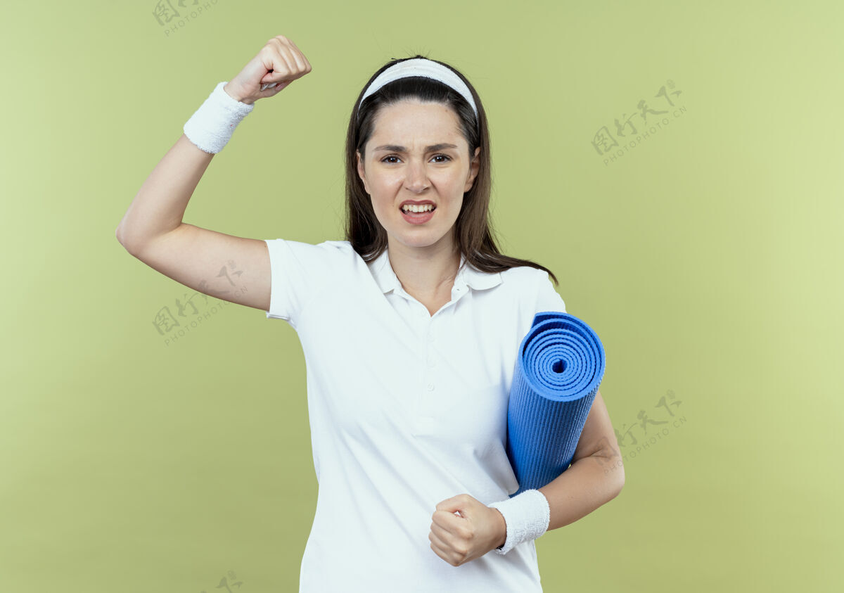 提高戴着头巾的年轻健身女士举着瑜伽垫举起拳头 怒气冲冲地站在轻墙上头巾拳头轻