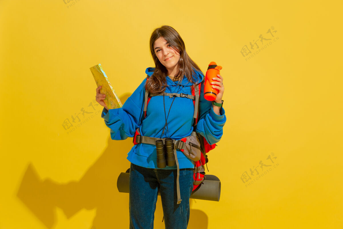 欢呼黄色摄影棚墙上 一个带着包和望远镜的快乐的年轻旅游女孩的画像人快乐女人