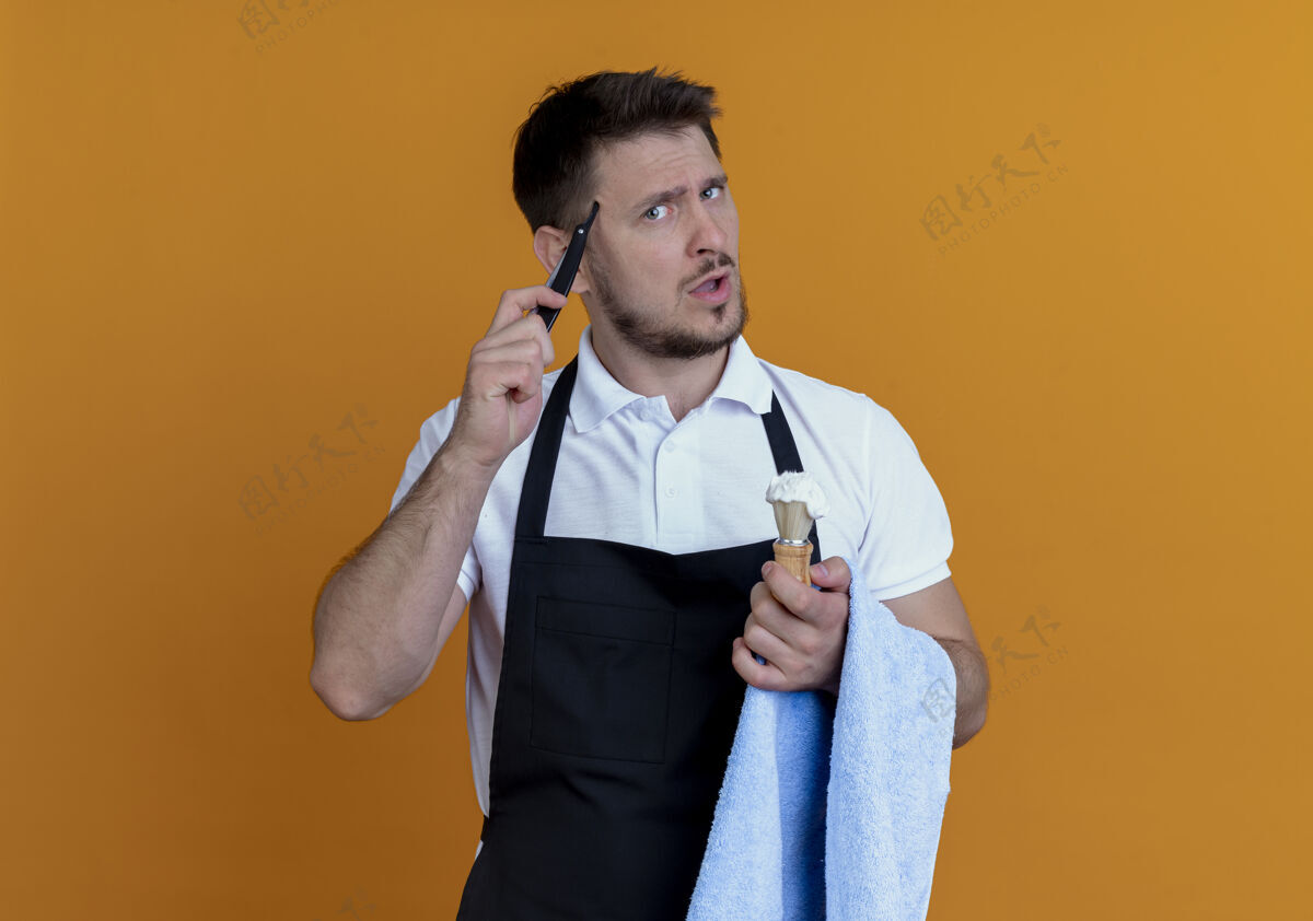 理发师围裙上的理发师手拿毛巾 手里拿着泡沫剃须刷和剃须刀 站在橙色的墙上 困惑不解剃须刀刷子围裙