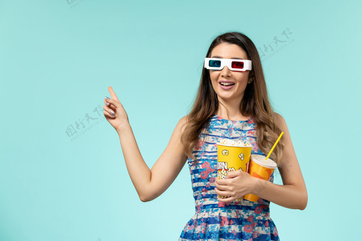 漂亮正面图年轻的女性拿着爆米花 戴着d墨镜在蓝色的表面上喝酒爆米花年轻的女性太阳镜