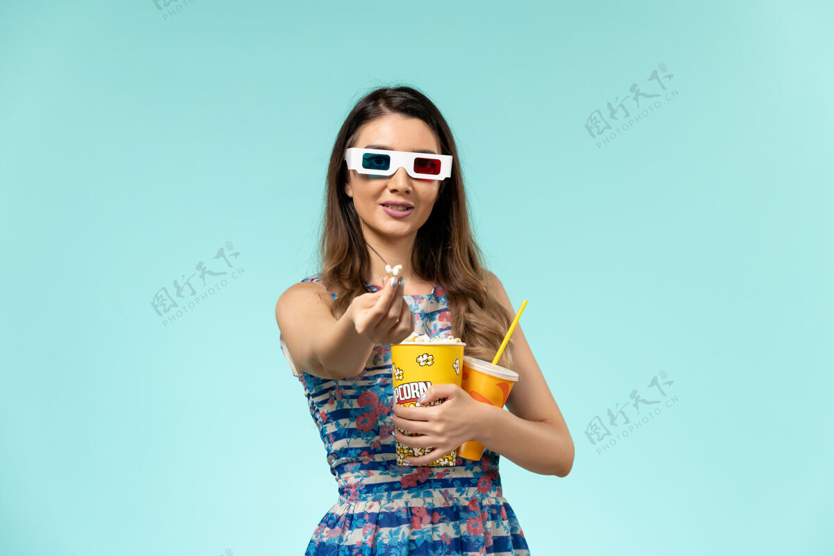 持有正面图年轻女性手持爆米花包 戴着d墨镜在浅蓝色表面喝酒年轻的女性剧院电影院