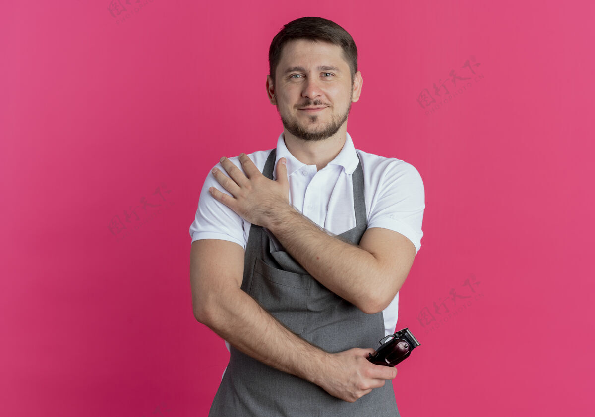 男人围裙上的理发师拿着剃须刀 自信地看着站在粉红色墙上的人修剪者理发师抱着