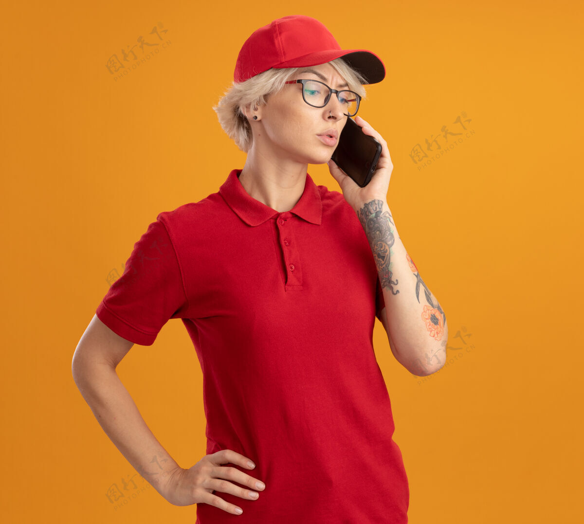 年轻身穿红色制服 戴着眼镜的年轻送货员站在橙色的墙上 严肃地讲着手机帽子脸站着