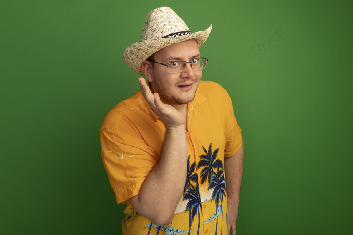 帽子戴着眼镜的男人穿着橙色衬衫 戴着夏天的帽子 手靠近脸微笑着站在绿色的墙上脸戴着站着
