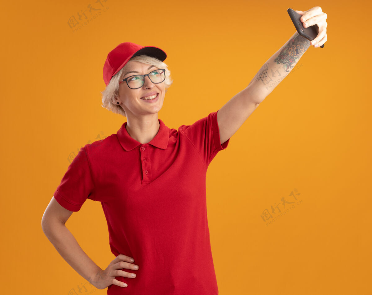 眼镜身穿红色制服 戴着眼镜的年轻送货员正在用智能手机自拍 站在橙色的墙上开心地微笑着制服站立女人