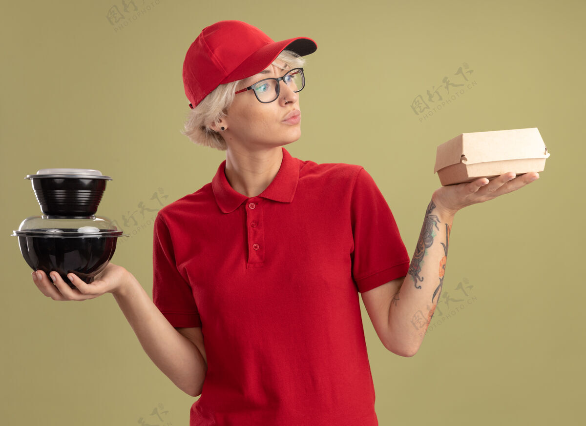 盒子年轻的送货员身穿红色制服 戴着帽子 戴着眼镜 手里拿着食品包 站在绿色的墙上 看起来很困惑 试图做出选择拿着女人试试