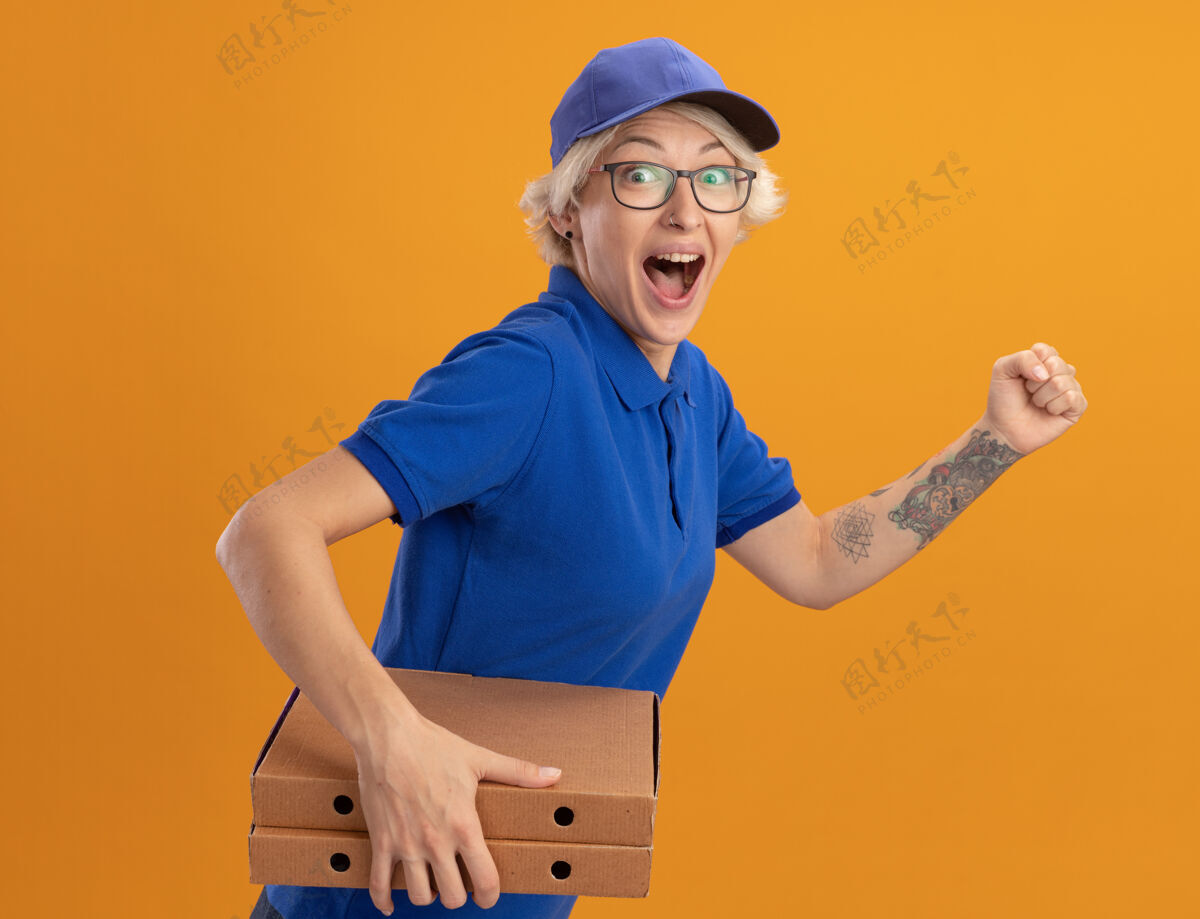 帽子身穿蓝色制服 头戴鸭舌帽的年轻送货员兴奋地跑过去 越过橘色的墙壁为顾客送比萨饼盒送货盒子顾客