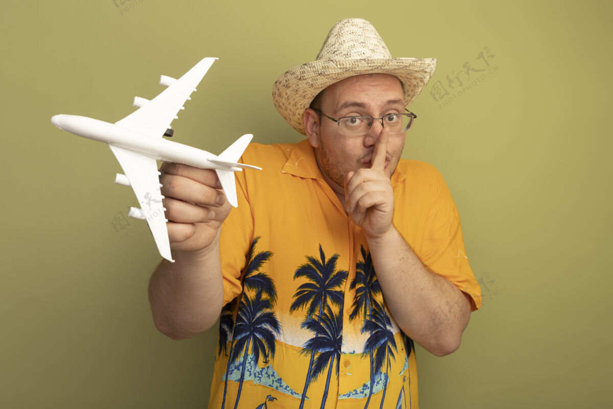 沉默戴着眼镜的男人穿着橘色衬衫戴着夏天的帽子拿着玩具飞机手指放在嘴唇上做着沉默的手势站在绿色的墙上手势男人站着