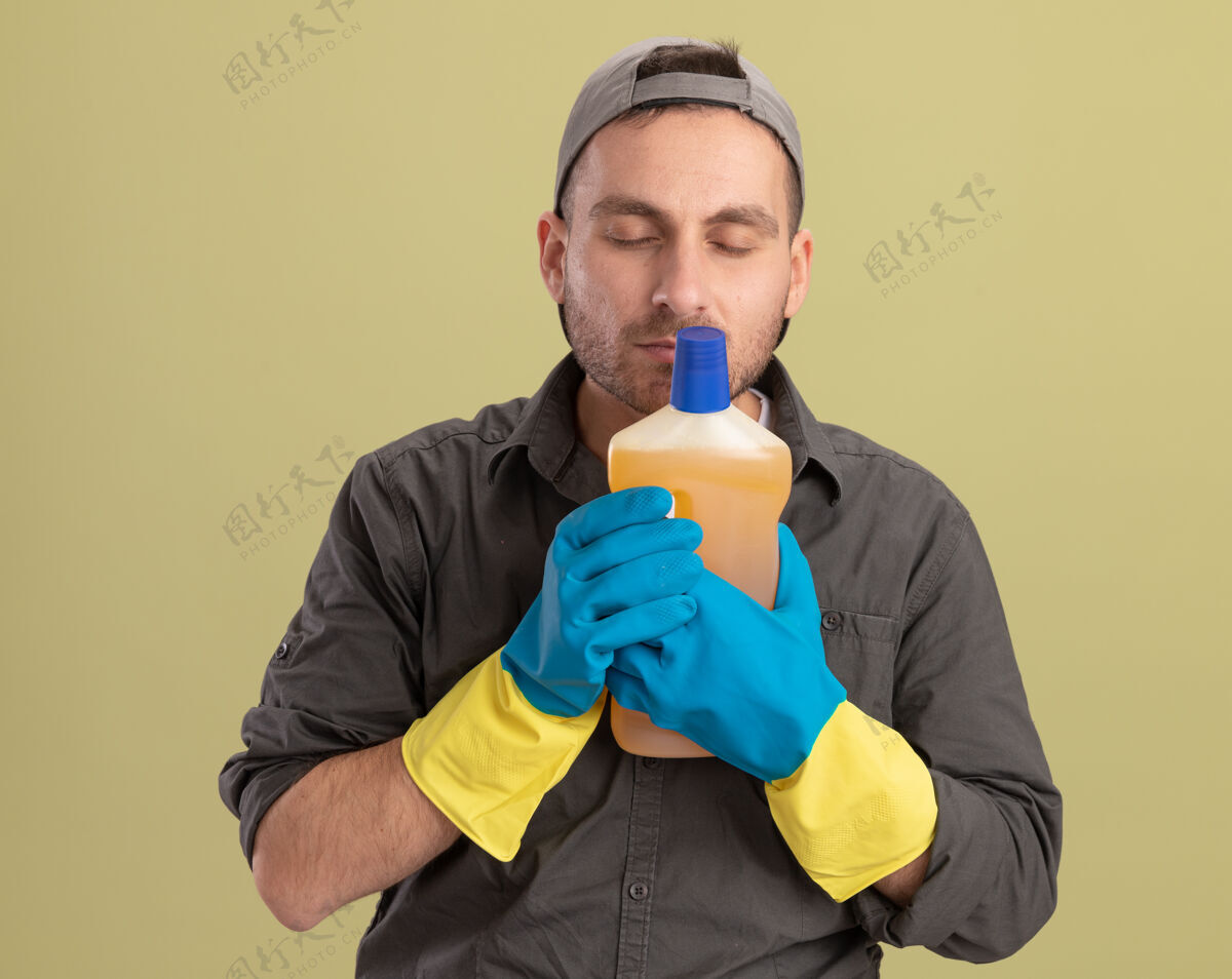 橡胶年轻的清洁工穿着休闲服 戴着橡胶手套 手里拿着一瓶清洁用品 呼吸着新鲜的香气 站在绿色的墙上手套新鲜瓶子
