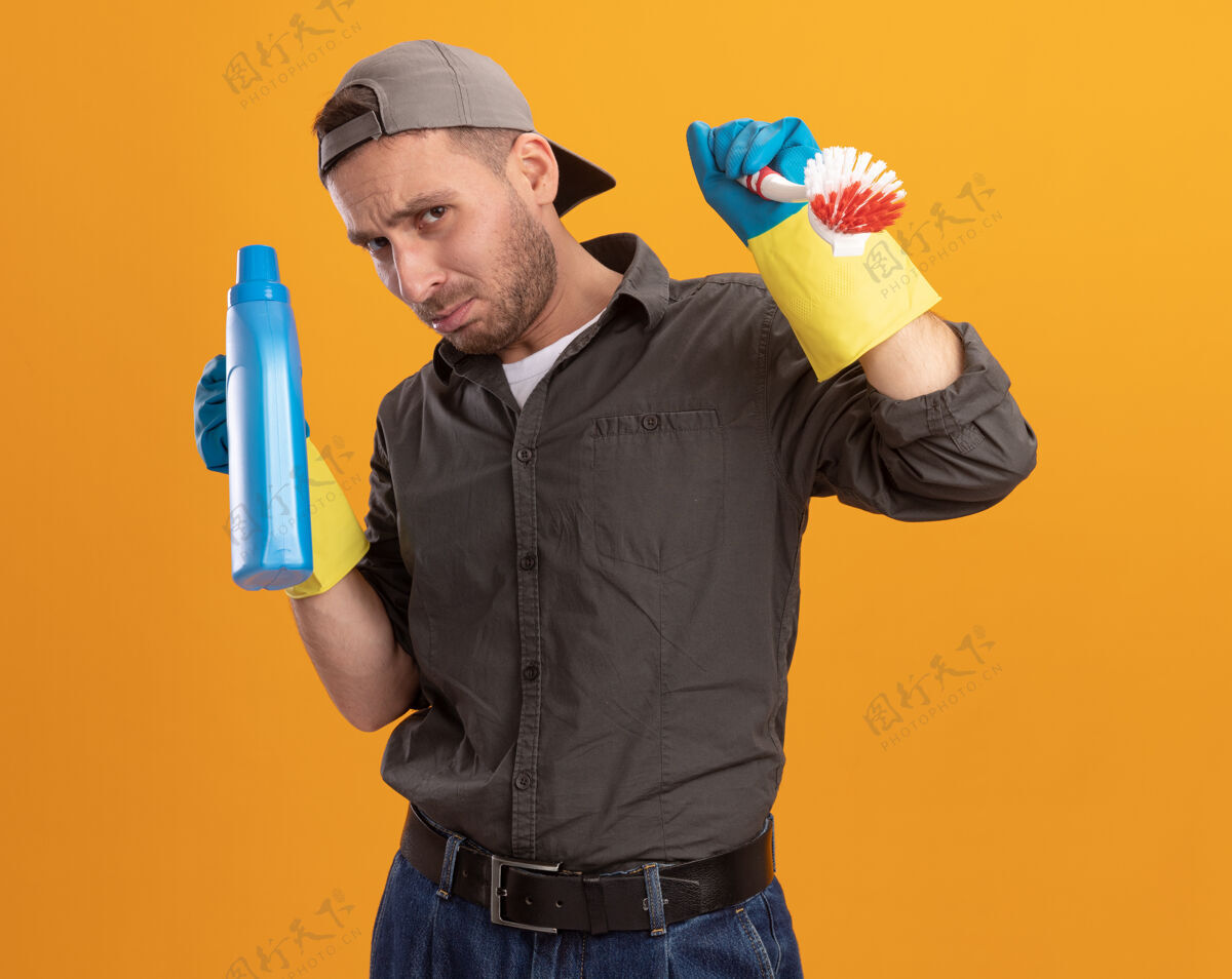 男人年轻的清洁工穿着休闲服 戴着橡胶手套 戴着帽子 手里拿着清洁刷和瓶子 手里拿着清洁用品 站在橘色的墙上 看上去既困惑又不高兴帽子刷子持有