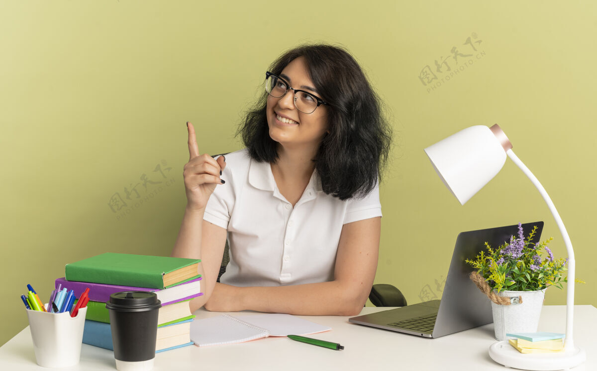 绿色年轻的笑容可掬的高加索女学生戴着眼镜坐在书桌旁 手里拿着学习用具 抬头看着隔离在绿色空间里的一面 还有复印空间看复制微笑