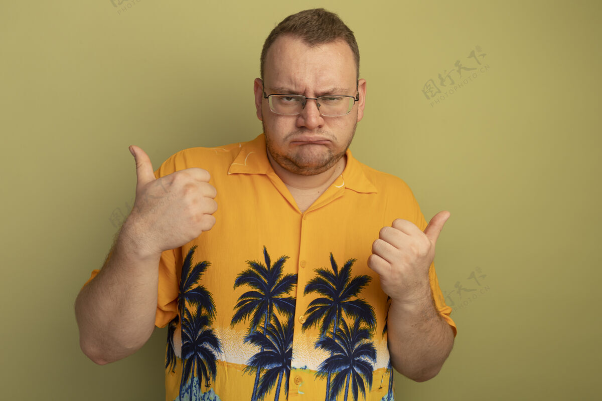 站一个戴着眼镜的男人 穿着橙色衬衫 怒气冲冲地皱着眉头 竖起大拇指站在明亮的墙上皱眉眼镜光