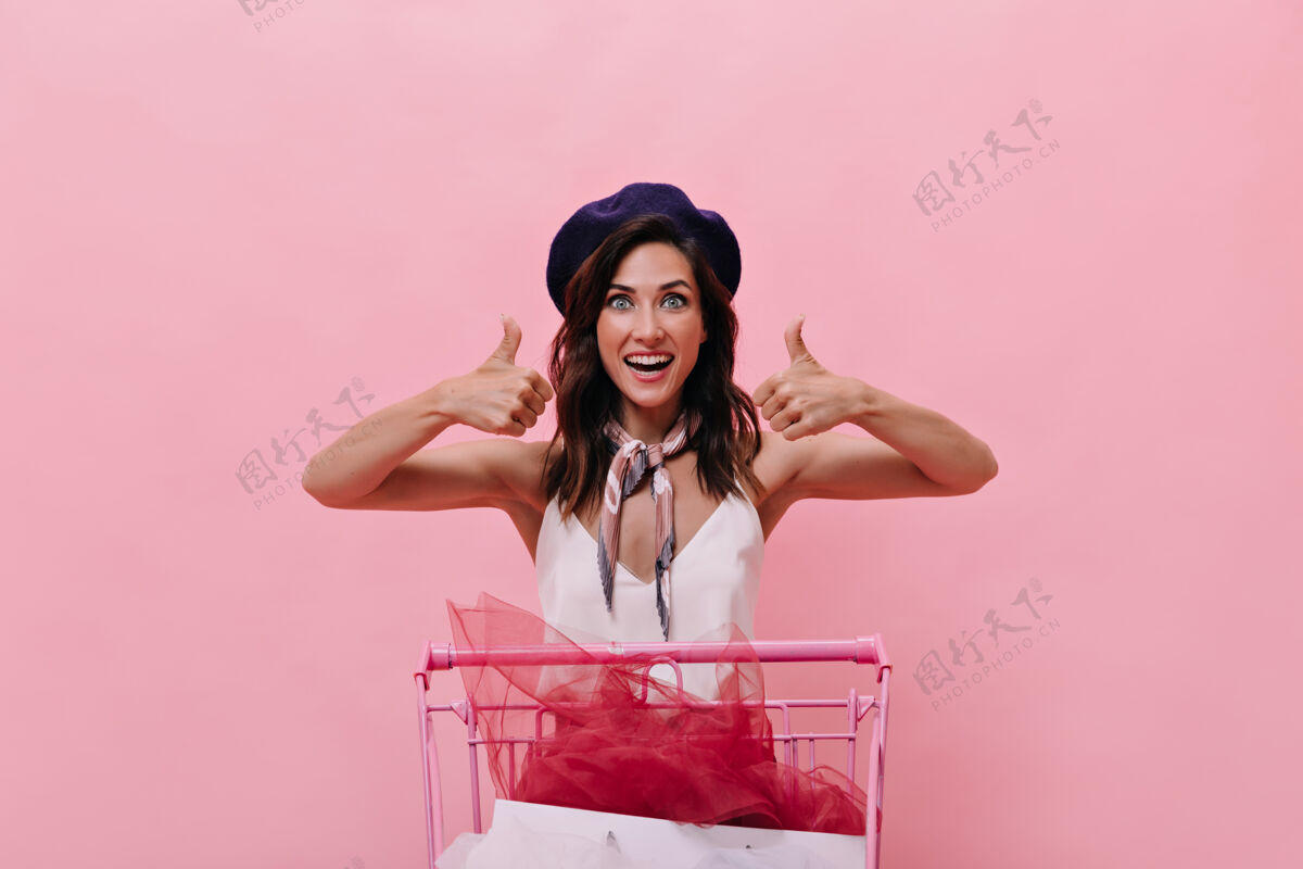肖像身穿蓝色贝雷帽的黑发女孩购物愉快 竖起大拇指心情愉快的女士戴着紫色帽子和白色衬衫 在粉色背景下摆姿势乐趣Swoosh健康