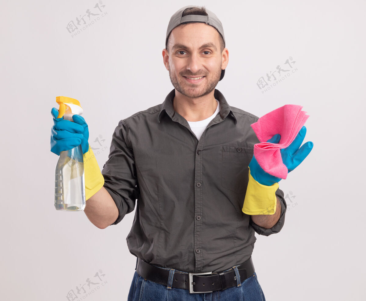 举行年轻的清洁工穿着休闲服 戴着橡胶手套 戴着帽子 手里拿着喷雾瓶和抹布 站在白色的墙上 脸上洋溢着幸福和积极的微笑男人抹布立场