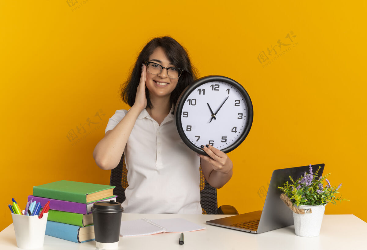 手年轻的笑容可掬的白人女学生戴着眼镜坐在书桌旁 手放在脸上 拿着时钟隔离在橙色空间和复印空间女学生坐拿