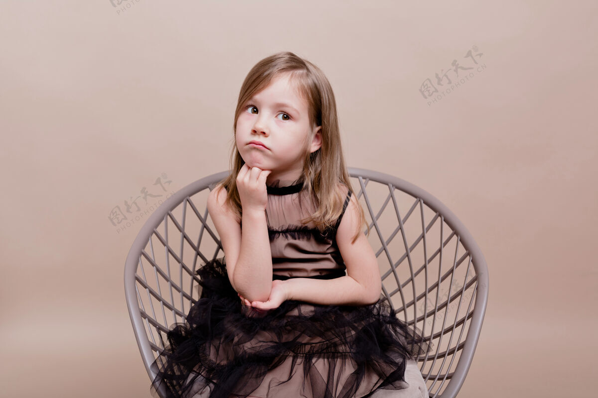快乐可爱的小女孩穿着黑色连衣裙坐在椅子上 一脸沉思 在孤零零的墙上摆着姿势 真是一个严肃的动作 漂亮迷人的女孩爱年轻魅力