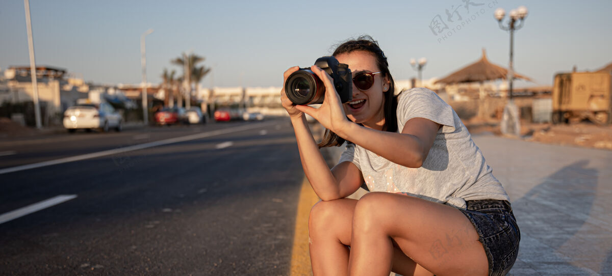 度假夏天 一个穿着短裤和t恤衫的年轻女子拿着专业相机坐在人行道上坐着散步日落