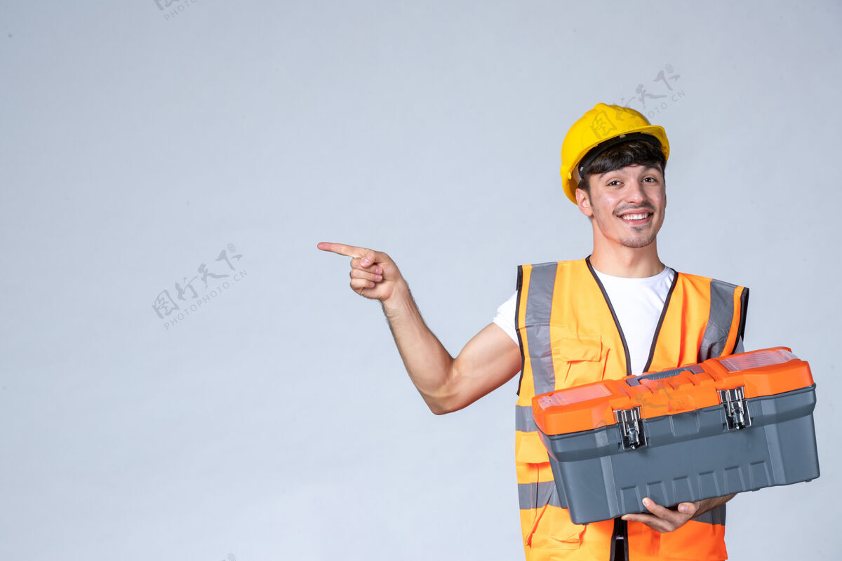 承包商正面图：年轻的男工人拿着沉重的工具箱 背景是白色的白人工作建筑工
