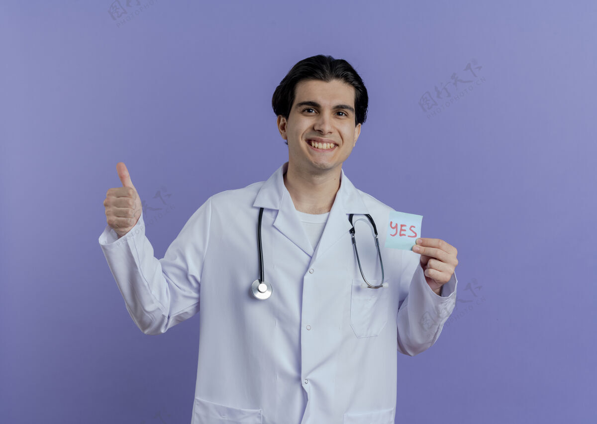 看快乐的年轻男医生穿着医用长袍 听诊器显示是的 注意到拇指向上 隔离在紫色的墙上 留有复印空间医生听诊器医疗