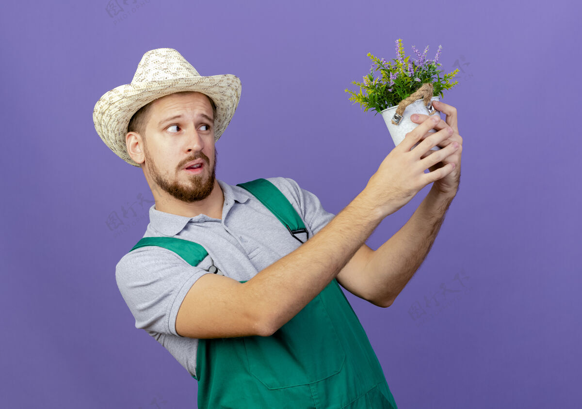 帽子年轻帅气的斯拉夫园丁穿着制服 戴着帽子 望着紫色墙上孤立的花盆花盆拿着英俊