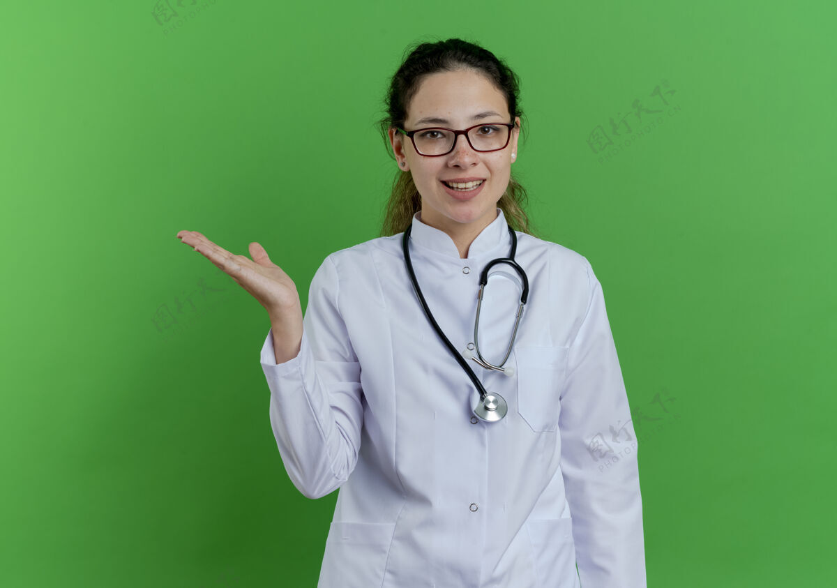 医生快乐的年轻女医生 穿着医用长袍 戴着听诊器和眼镜 在绿色的墙壁上展示着被隔离的空手和复印空间女长袍空