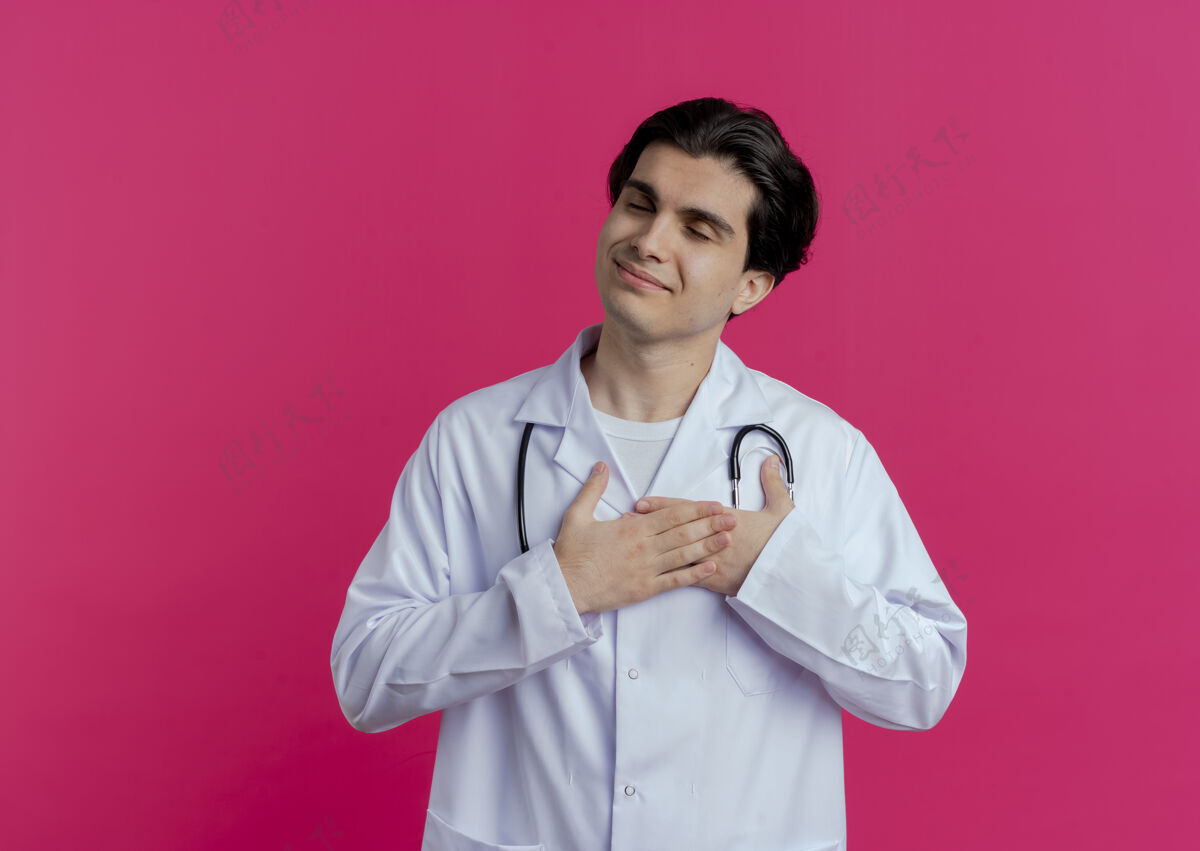 保存高兴的年轻男医生穿着医用长袍和听诊器 双手放在心上 闭着眼睛 隔离在粉红色的墙上 留有复印空间医生听诊器封闭