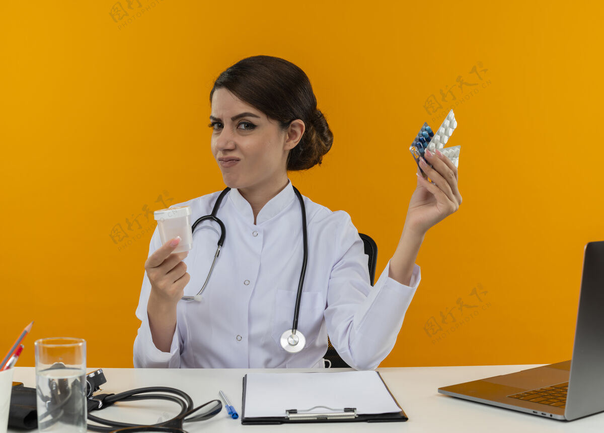 工具穿着医用长袍和听诊器的年轻女医生坐在桌子旁 手里拿着医疗工具和手提电脑 拿着隔离在黄色墙上的药品和烧杯穿烧杯医疗