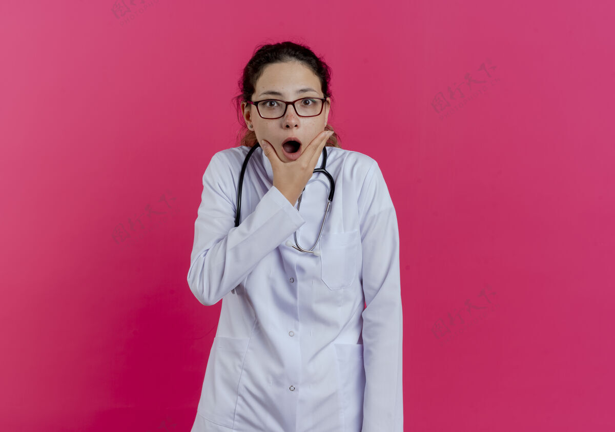 医生年轻的女医生穿着医用长袍 戴着听诊器 戴着眼镜 把下巴隔离在粉红色的墙上 留着复印空间看下巴女