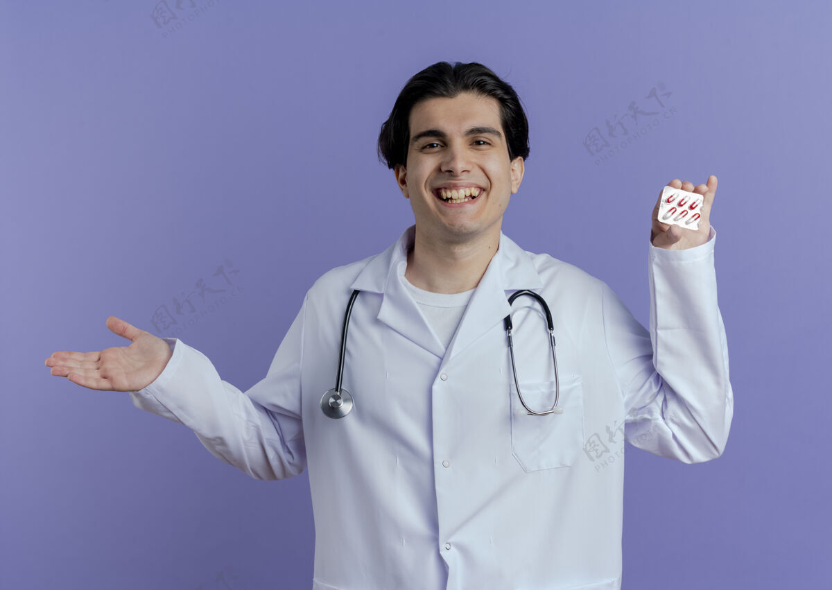 胶囊快乐的年轻男医生穿着医用长袍和听诊器 在紫色的墙上展示一包医用胶囊和一只空手空表演年轻