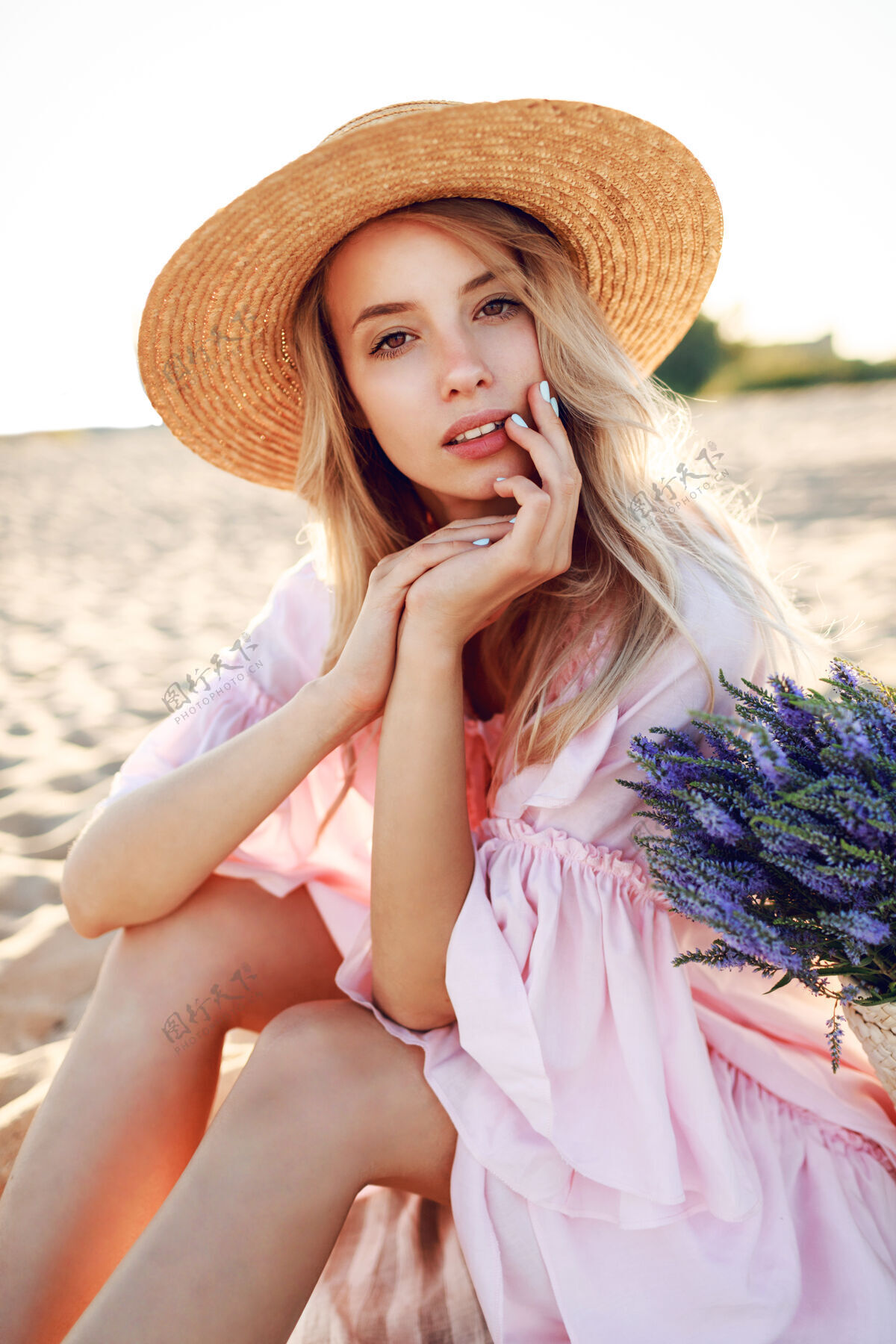 健康浪漫的白人女子 戴着时髦的帽子 穿着优雅的粉色连衣裙 手持草包和花束 在沙滩上摆着姿势自由看起来时尚
