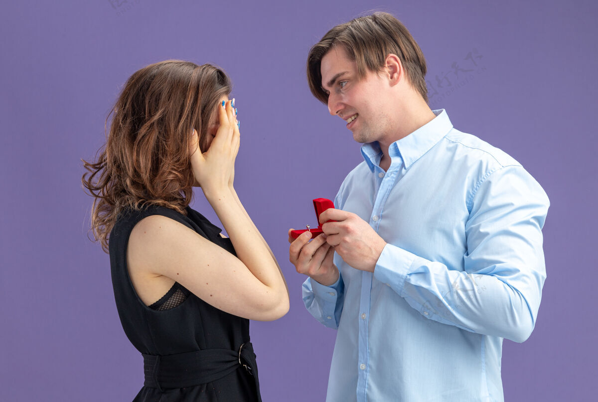 戒指一对年轻漂亮的情侣幸福地戴着红盒子里的订婚戒指 站在蓝色的背景上 向情人节用手捂住眼睛的可爱女友求婚男人女朋友制造