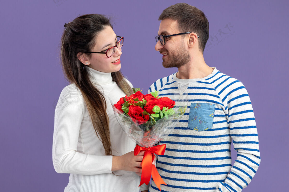 情人节一对年轻漂亮的情侣穿着休闲服 手持一束红玫瑰的快乐女人站在紫色的墙上 看着微笑着庆祝情人节的男友庆祝情侣男朋友