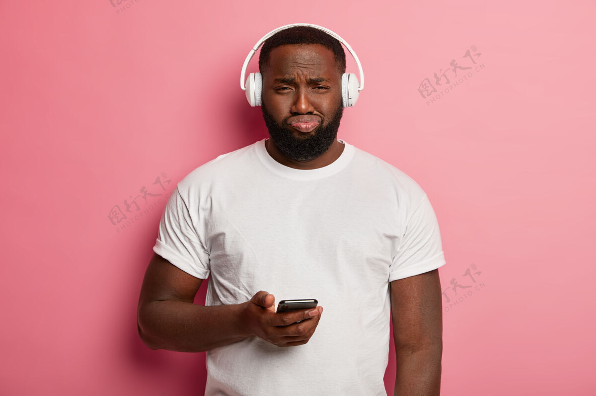 歌曲不高兴的黑人没刮胡子的男人戴着耳机听音乐 有不高兴的表情 拿着手机 穿着随便 心烦意乱得无法在播放列表中下载歌曲不高兴拍不高兴