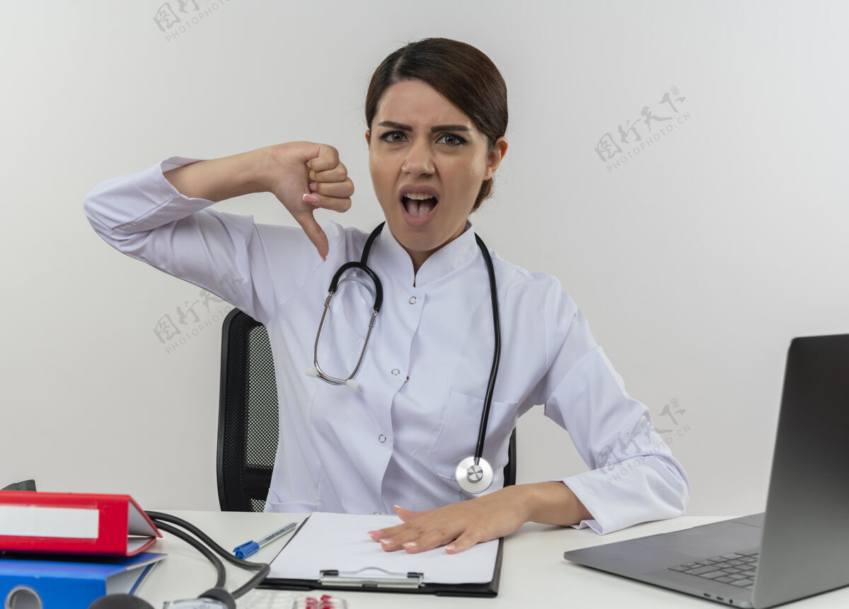 穿恼怒的年轻女医生穿着医用长袍和听诊器坐在办公桌旁 手里拿着医疗工具和笔记本电脑 拇指朝下 被隔离在白墙上拇指女医疗