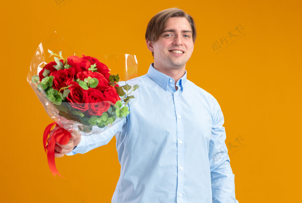 微笑身着蓝衣的快乐男人手持一束红玫瑰 看着相机 站在橙色的墙上 欢快地微笑着情人节的概念概念玫瑰日子