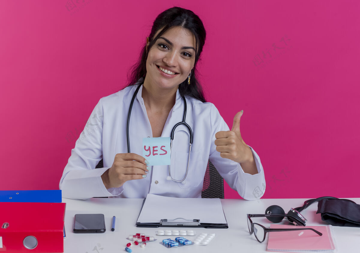 表演微笑着的年轻女医生穿着医用长袍和听诊器坐在办公桌旁 手里拿着医疗工具 手里拿着一张“是”的纸条 在粉红色的墙上孤立地竖起大拇指穿拇指医生