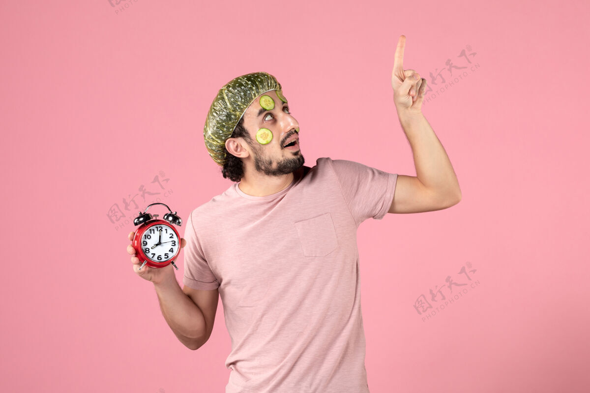 光滑正面图：戴着面具的年轻男性手持粉色背景的时钟皮肤年轻男性视图