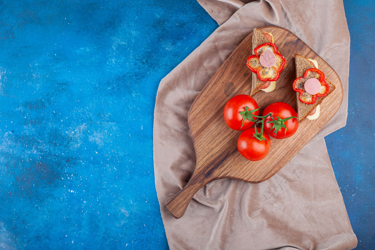 面包三明治上放着香肠和整个西红柿 放在切菜板上 蓝色的布料上酵母美味美味