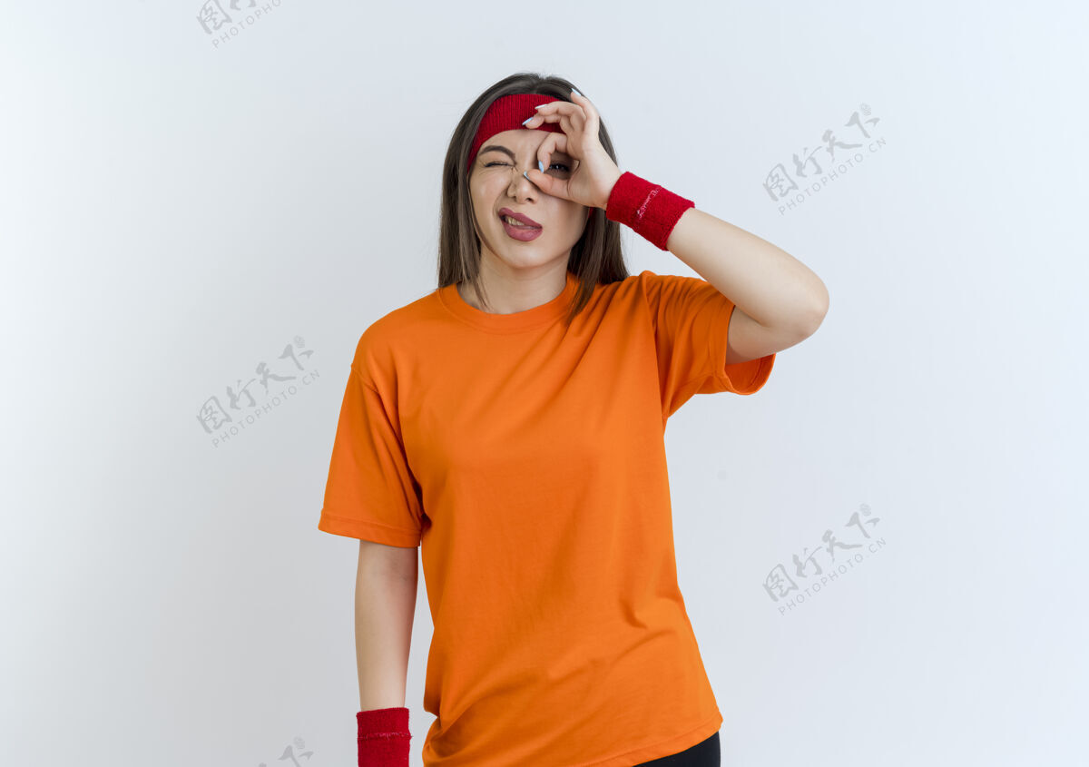 眨眼自信的年轻运动女性戴着头带和腕带做造型手势眨眼隔离在白色墙壁与复制空间女人表情腕带