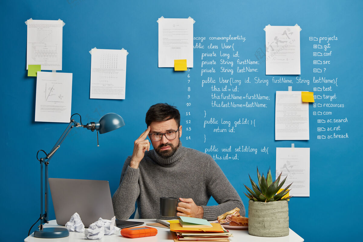 男性戴眼镜的胡子男人在思考创业计划 有不愉快的表情 努力集中精神 喝咖啡 在自己的柜子里做些遥远的工作笔记笔记本电脑人