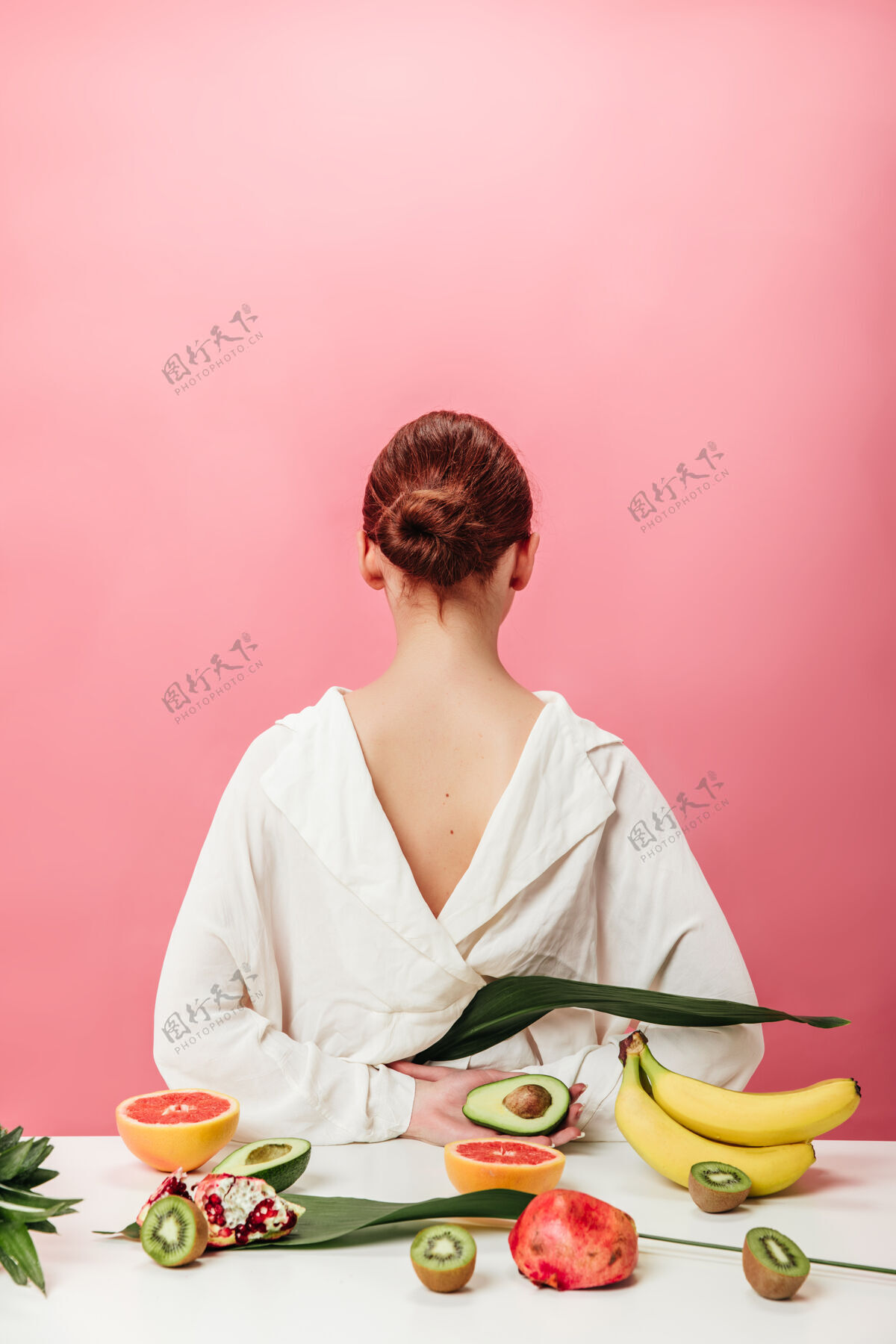 年轻人姜味女人的后视图和异国水果摄影棚拍摄的女孩在白色衬衫与香蕉 葡萄柚 石榴石猕猴桃和鳄梨粉红色的背景粉色石榴食物