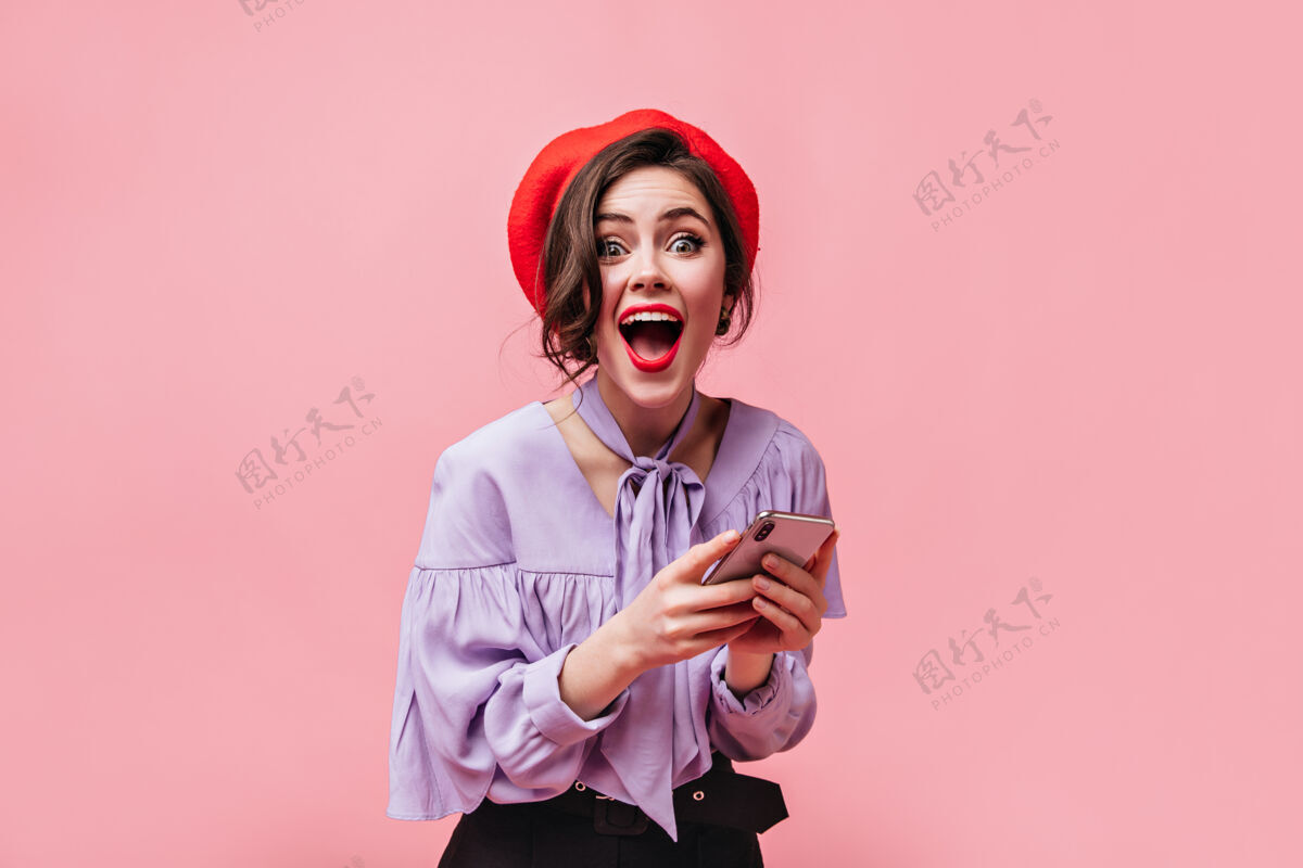 黑色身着红色贝雷帽和时尚衬衫的情绪化女人在惊喜中看着相机 手持粉色背景的白色智能手机肖像微笑裤子