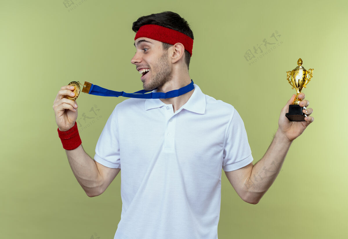 快乐戴着头巾 脖子上戴着金牌的年轻运动型男子手持奖杯 为绿色而高兴和兴奋金兴奋抱