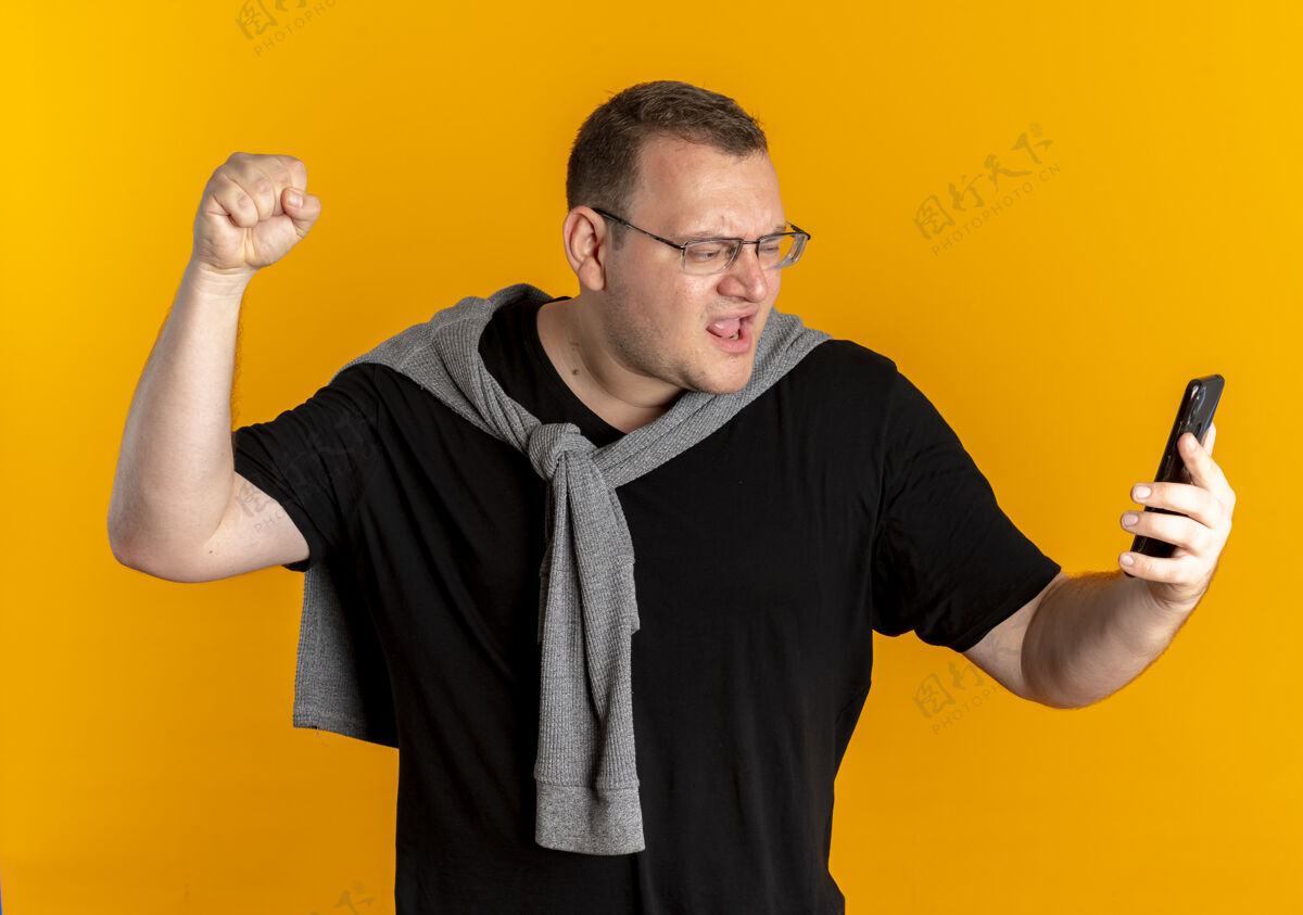 站着一个戴眼镜 身穿黑色t恤 手持智能手机的超重男子 握紧拳头 为自己战胜橘子而欢欣鼓舞手机戴着高兴
